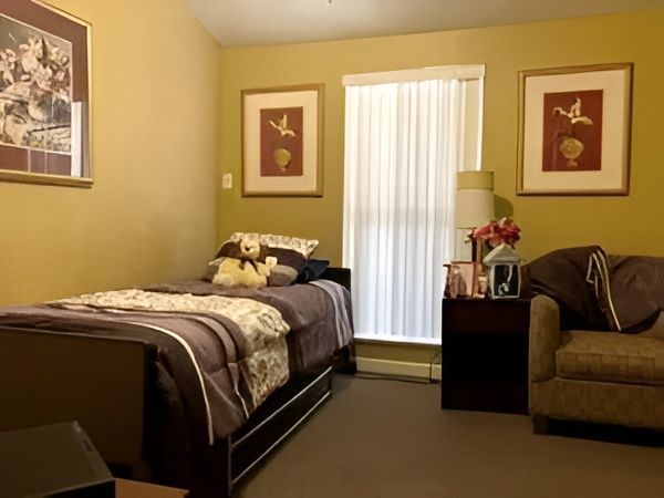 Neatly furnished bedroom with a bed, armchair, window, and framed wall art in warm beige tones.