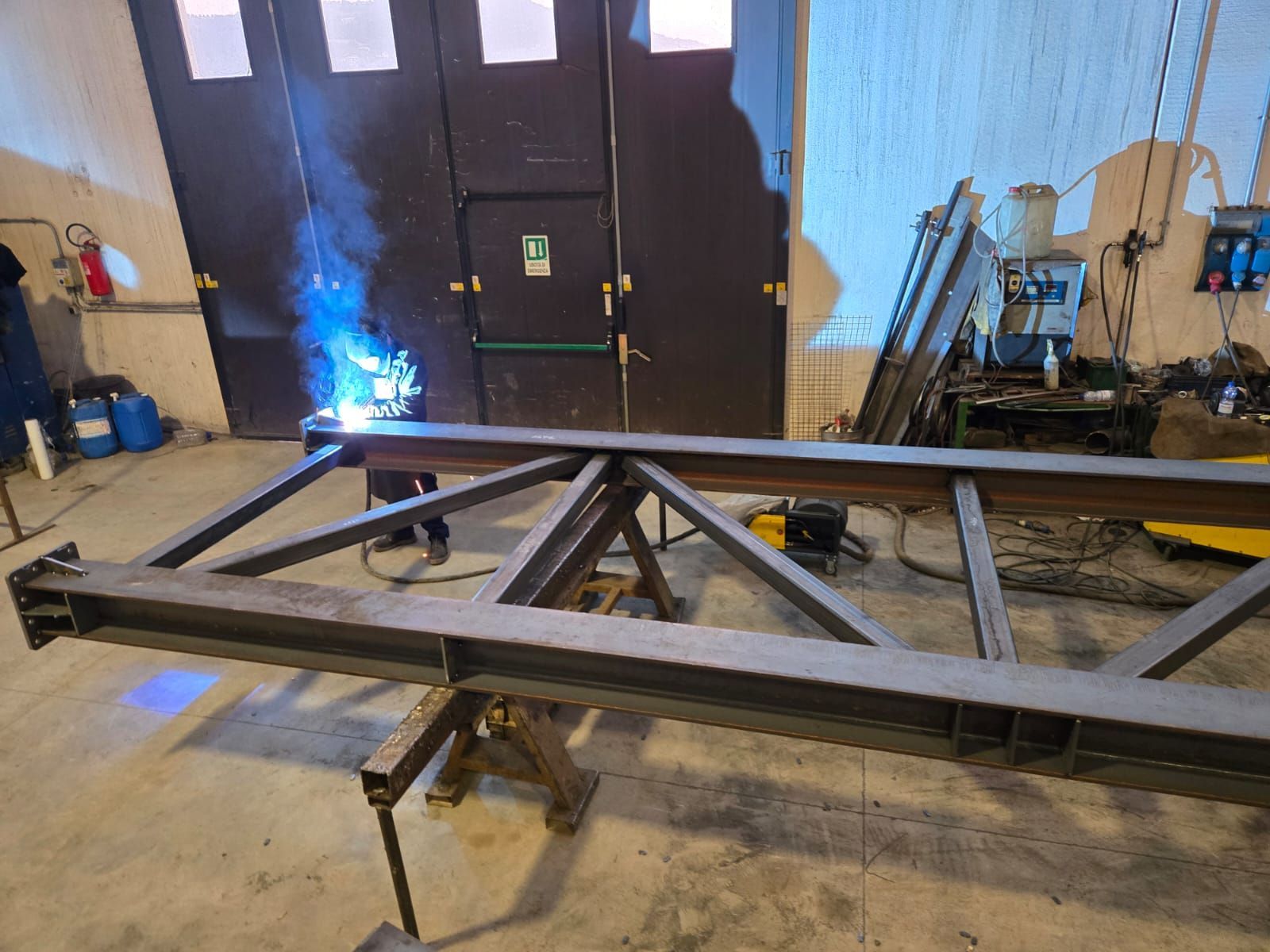 Un operaio utilizza una torcia per saldatura per unire travi metalliche sul pavimento di un'officina, producendo un arco blu brillante.