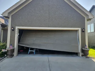 Damaged garage door, partially open. The door is bent and askew. A small stepladder is visible inside the garage.