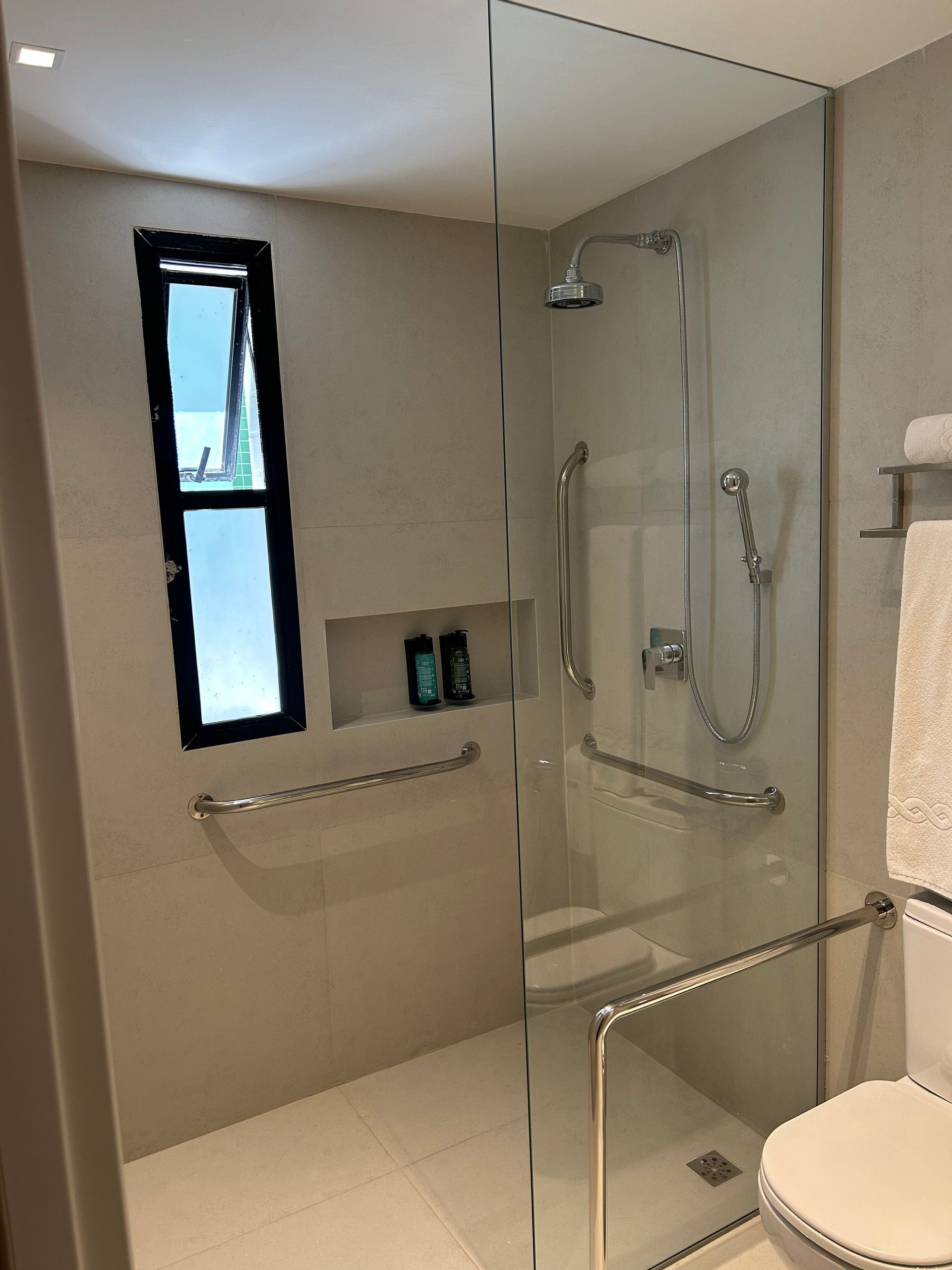 Banheiro acessível com chuveiro de acesso para cadeirantes, barras de apoio, banco dobrável e vaso sanitário parcialmente visível.