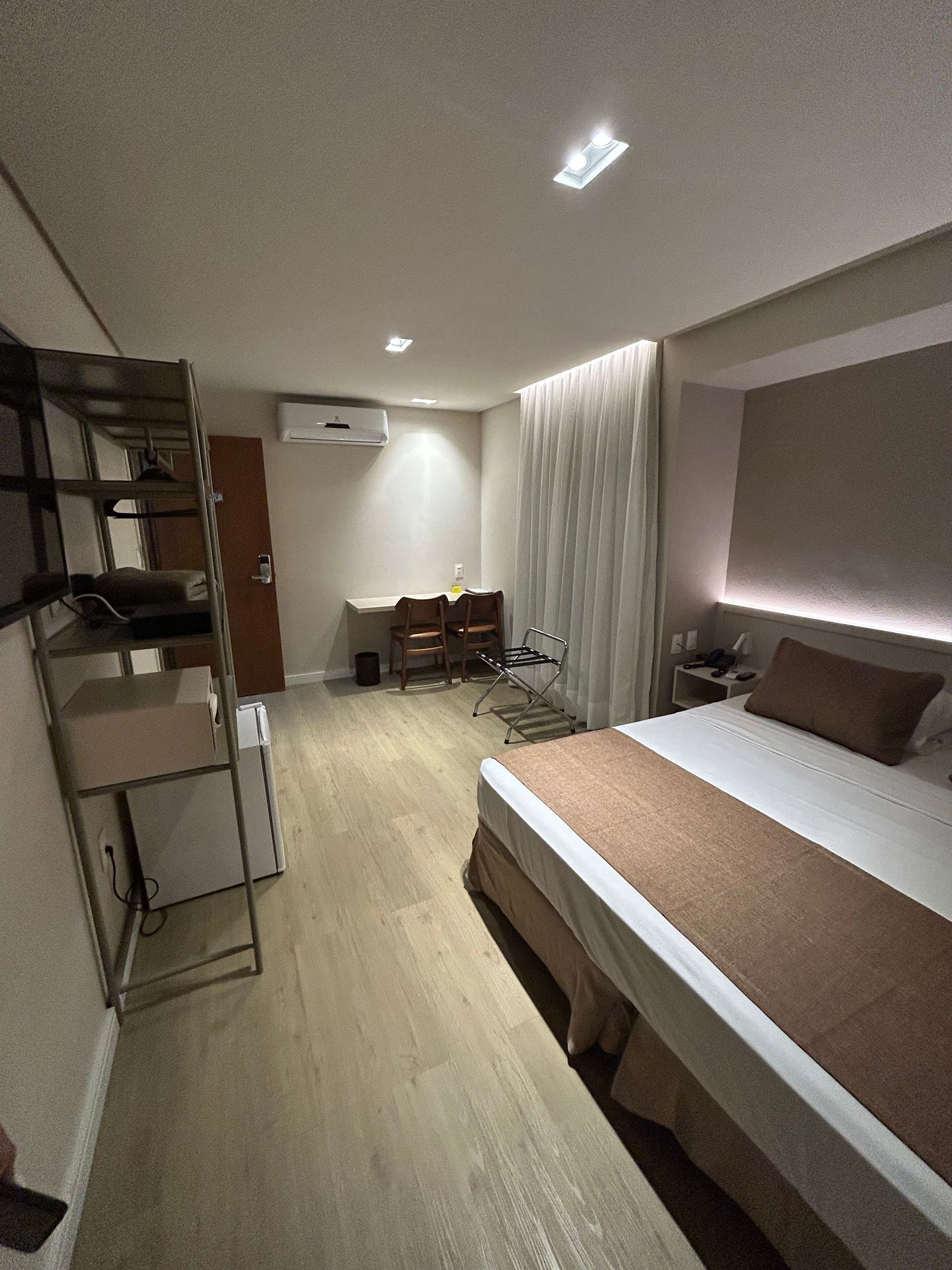 Quarto moderno com cama, prateleiras, escrivaninha e cortinas em tons neutros e aconchegantes.
