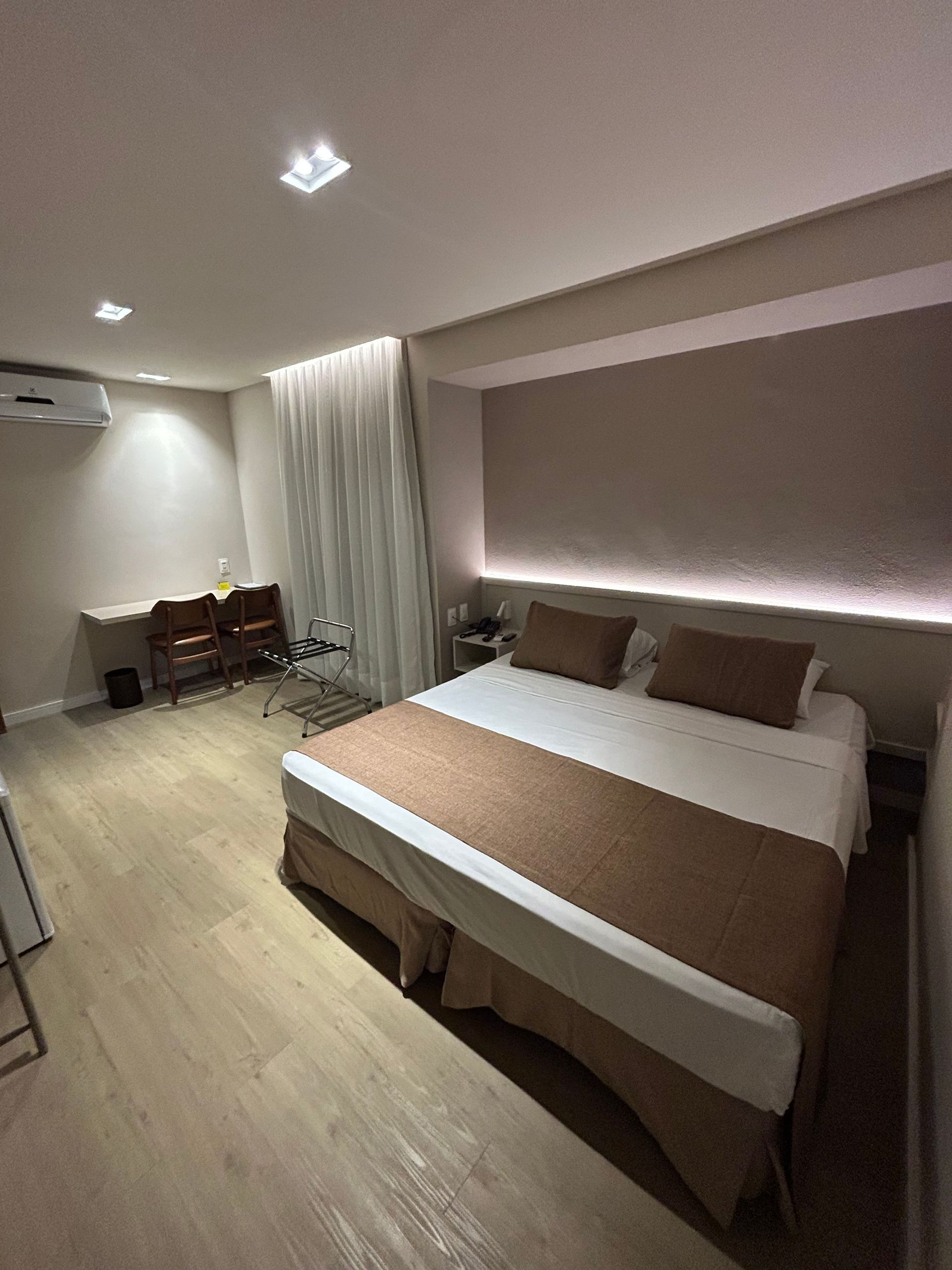 Quarto moderno com cama arrumada, piso de madeira, cortinas e iluminação suave na parede.