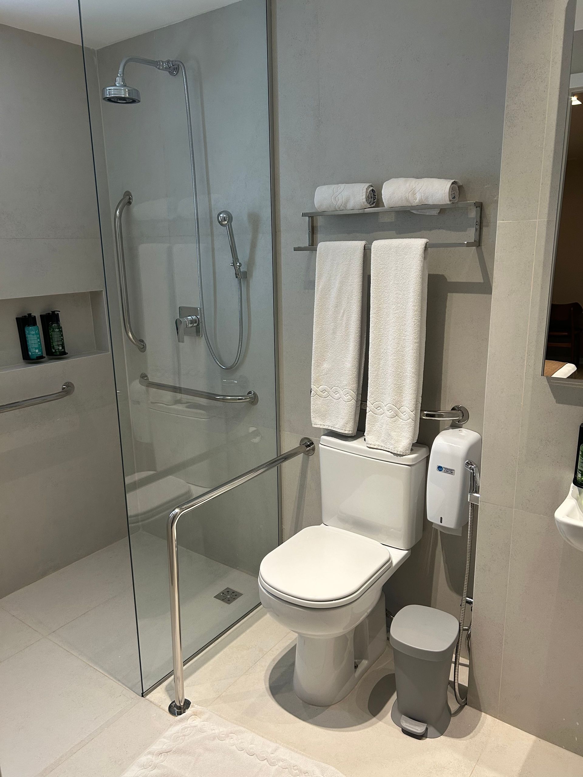 Banheiro acessível no hotel com chuveiro de acesso facilitado, vaso sanitário, pia e barras de apoio.
