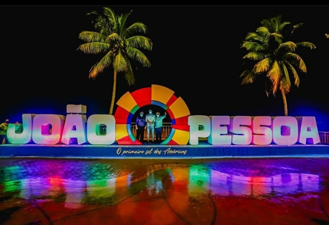 Letreiro colorido e iluminado com o nome “João Pessoa” à noite, com palmeiras e reflexos de arco-íris no chão molhado.