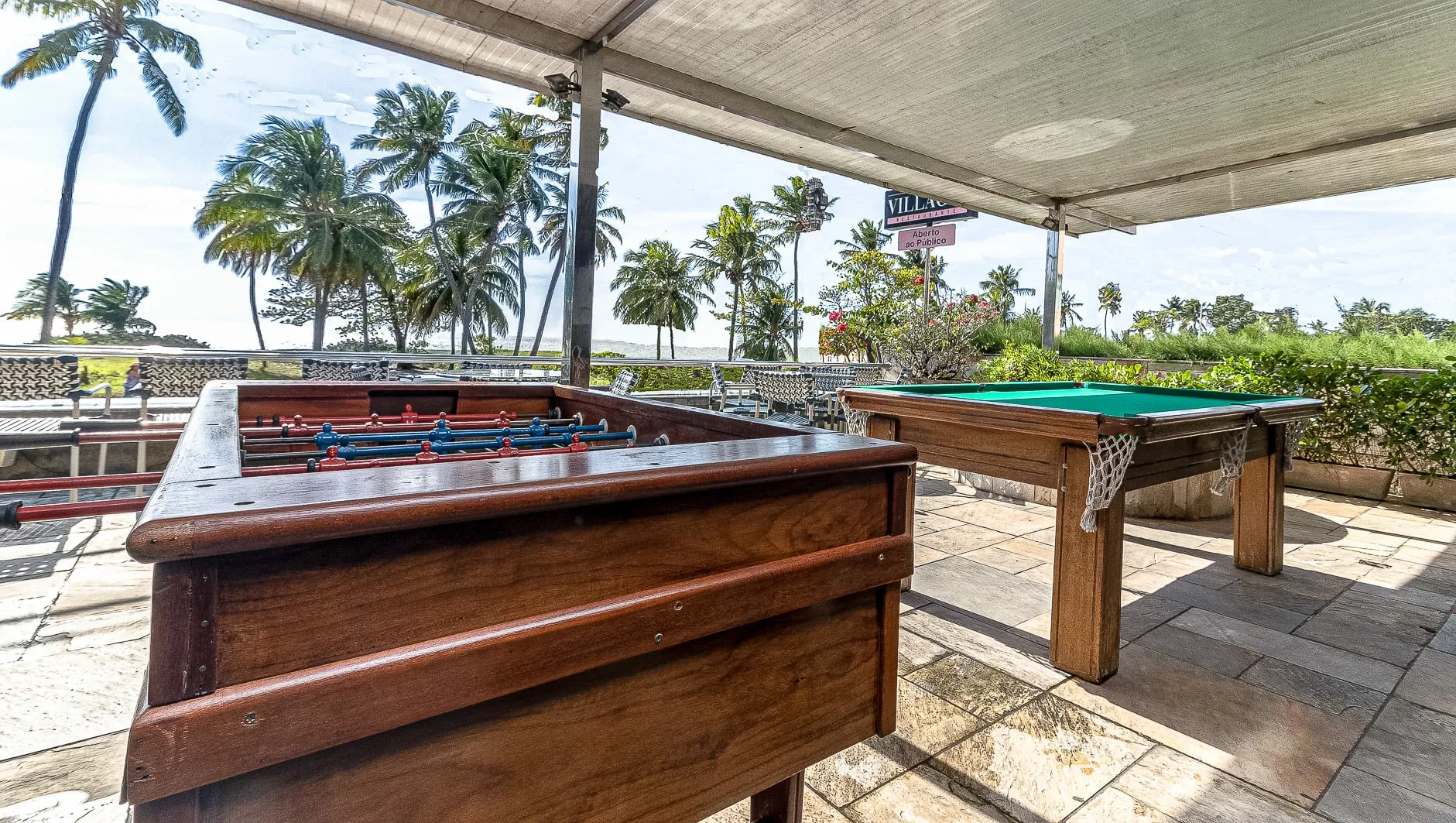 Área de jogos externa sombreada com mesa de bilhar de madeira em um pátio, palmeiras e o oceano ao fundo.
