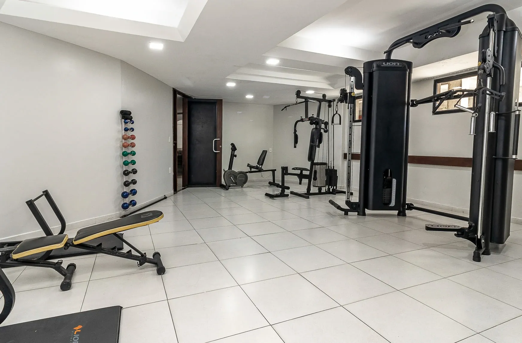 Academia moderna com aparelhos de musculação, banco de supino e piso de azulejo em uma sala branca e iluminada.