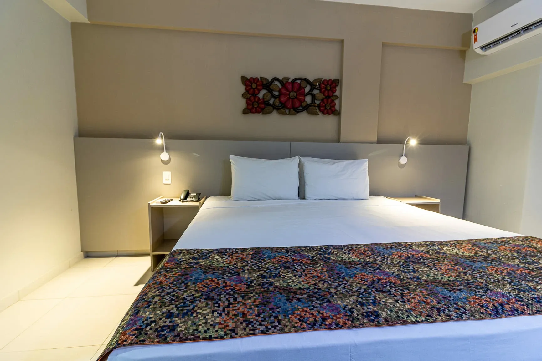 Quarto de hotel moderno com cama grande, tapete estampado, abajures e paredes bege.