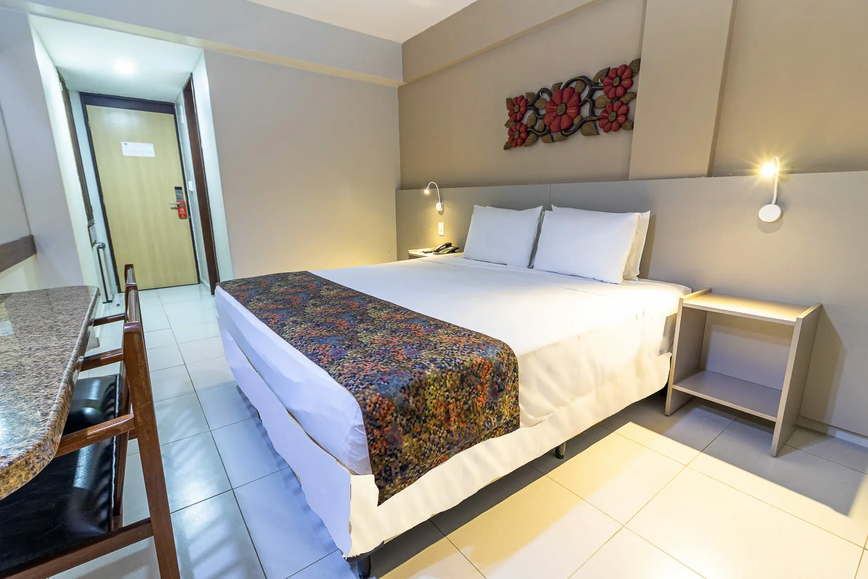 Quarto de hotel moderno com cama king-size, tapete estampado, abajures e piso de azulejo.