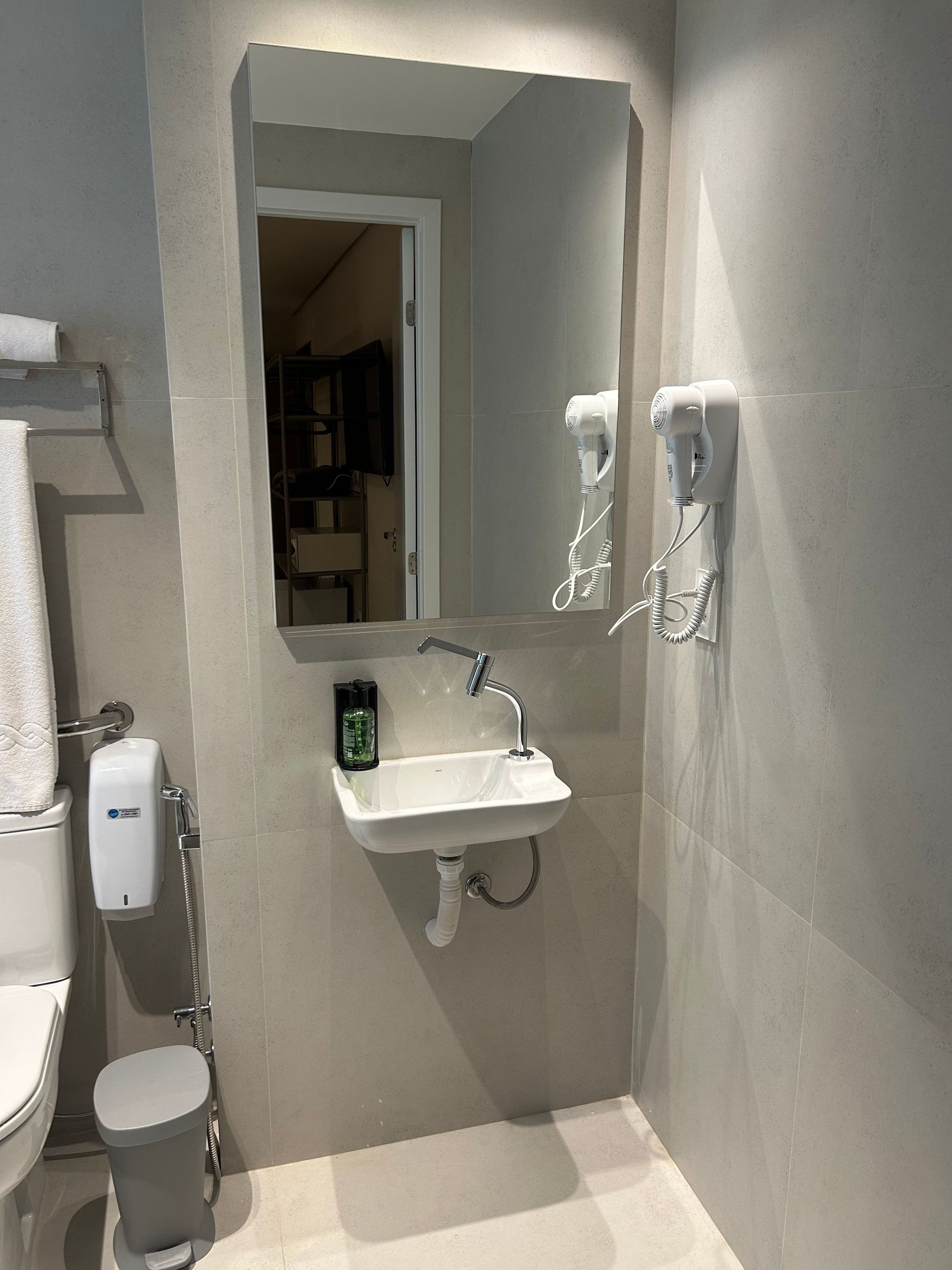 Pequeno banheiro acessível com pia, espelho, telefone fixado na parede e vaso sanitário visíveis à esquerda.