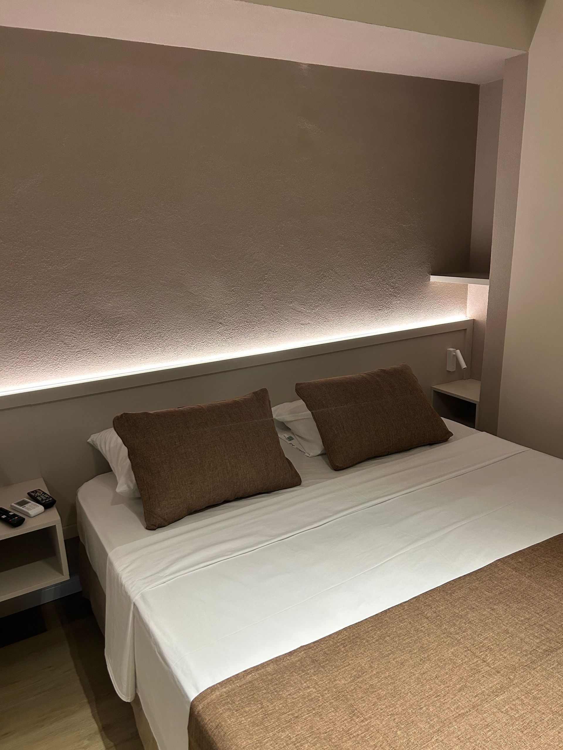Cama moderna estilo hotel com lençóis brancos, travesseiros marrons e cobertor bege sob um painel de parede iluminado.