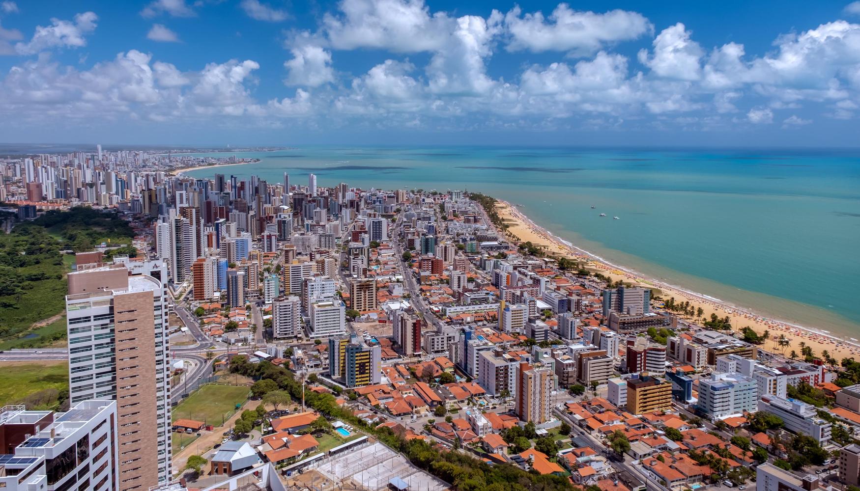 Vista aérea de uma cidade costeira com arranha-céus, praias e oceano turquesa sob um céu parcialmente nublado.
