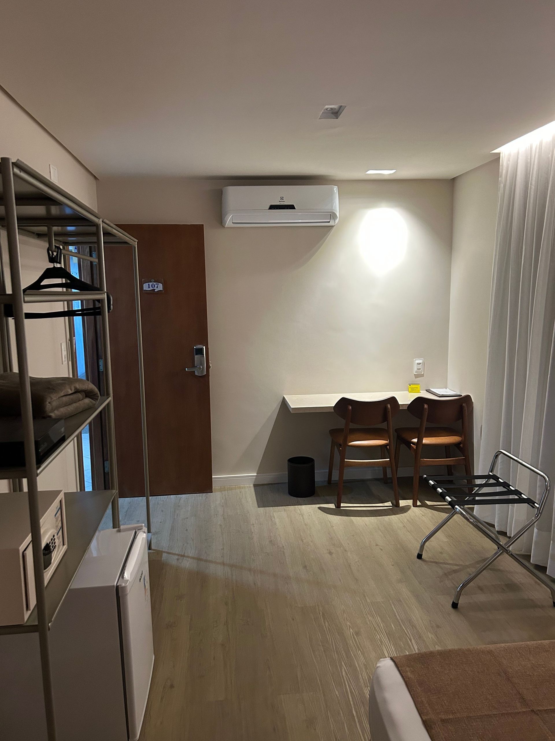 Quarto de hotel pequeno com escrivaninha, cadeiras, ar-condicionado, prateleiras e varal.