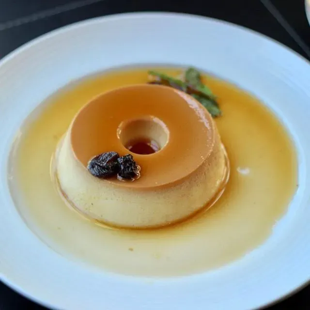 Sobremesa de caramelo estilo pudim servida em um prato branco, decorada com um pequeno raminho de hortelã e uma uva passa.