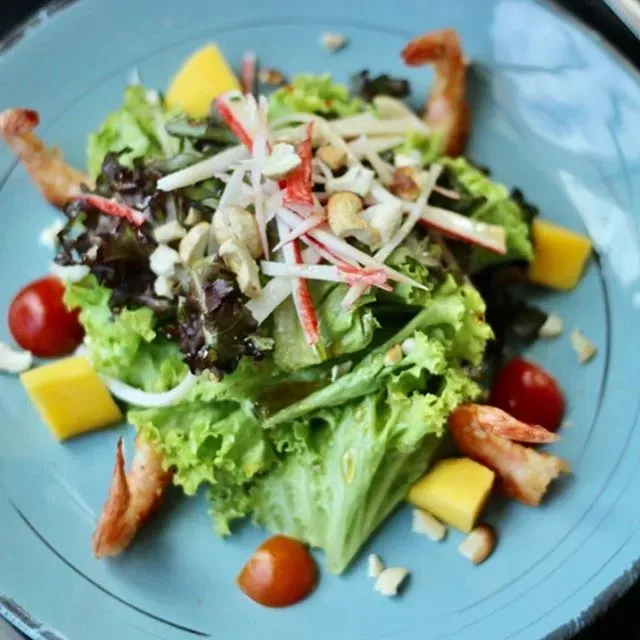 Salada fresca em uma tigela azul com alface, camarão, tomates-cereja, manga e kani desfiado.