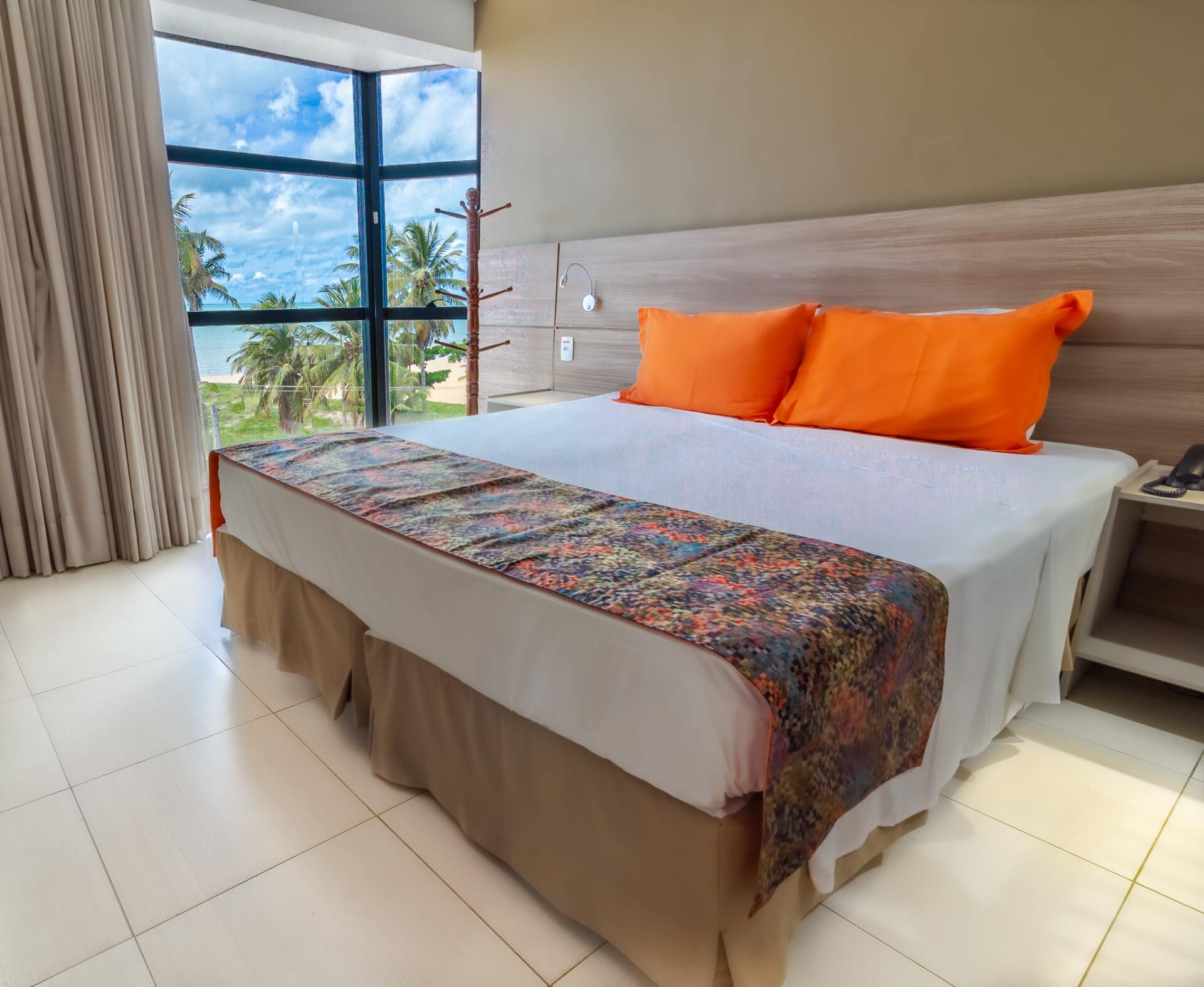 Quarto de hotel moderno com cama branca, travesseiros laranja, tapete estampado e ampla janela com vista para o mar.