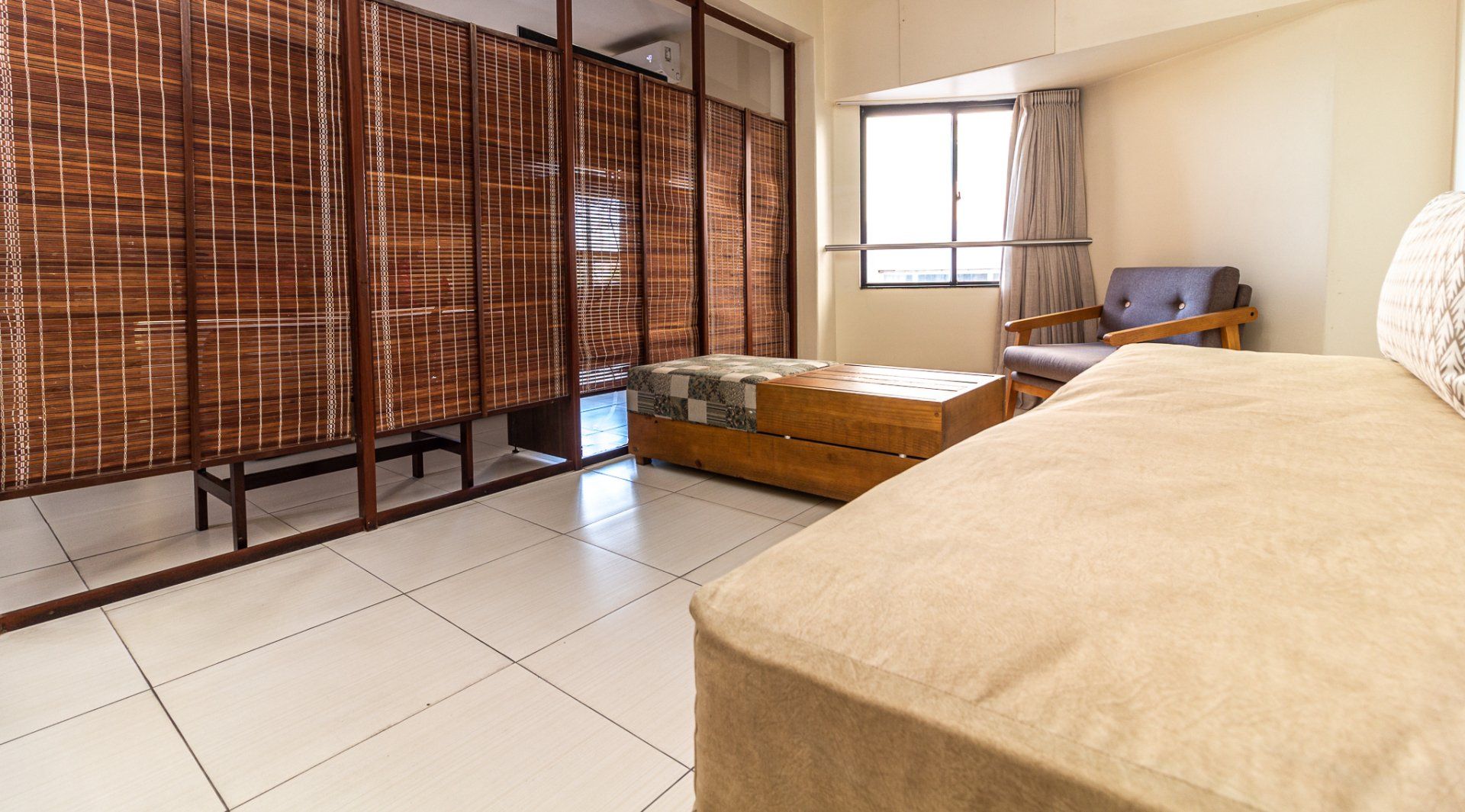 Quarto luminoso com divisória de bambu, cama baixa e recanto junto à janela, banhado por luz natural aconchegante.