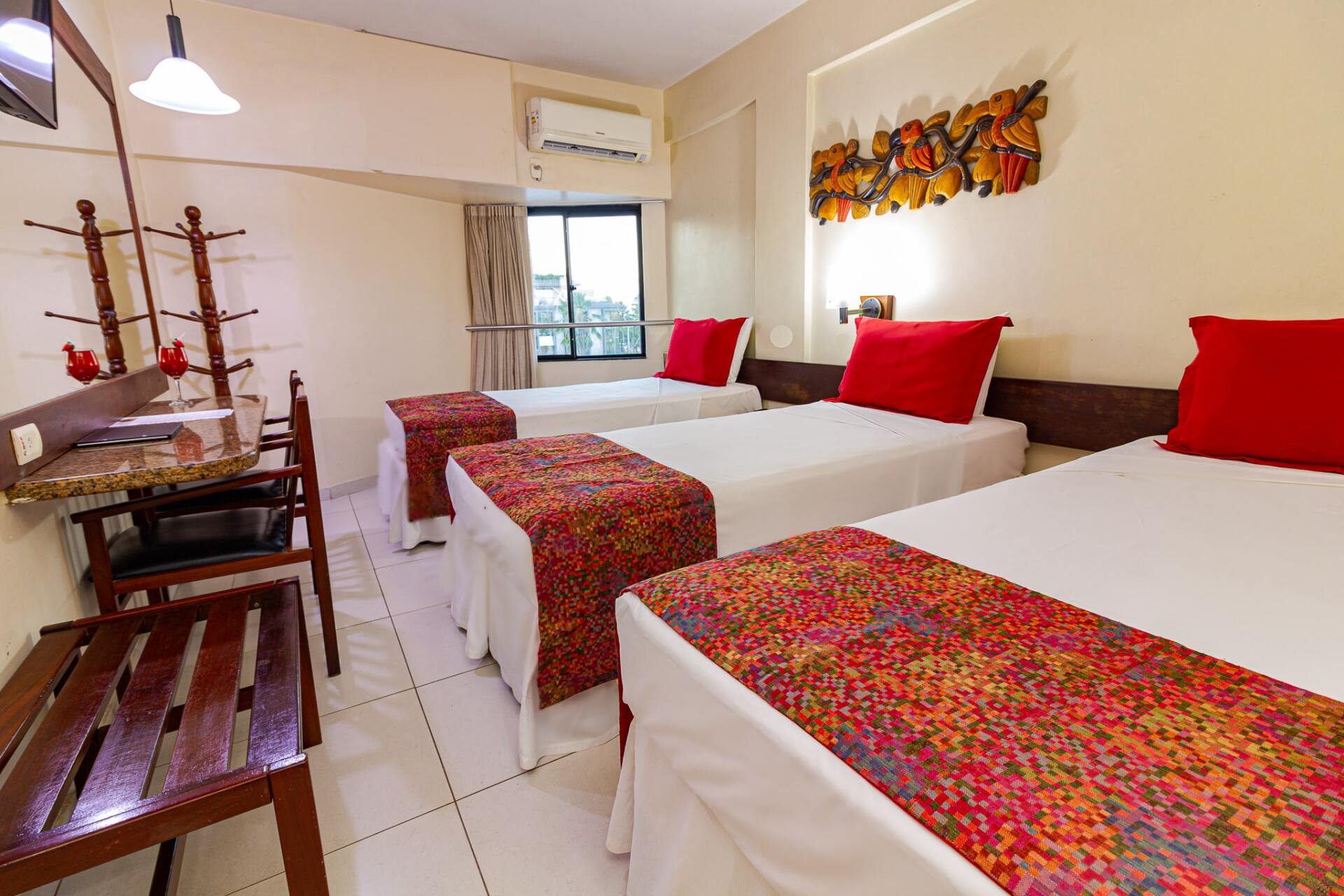 Quarto de hóspedes luminoso com três camas, detalhes em vermelho, cadeiras de madeira e uma janela.