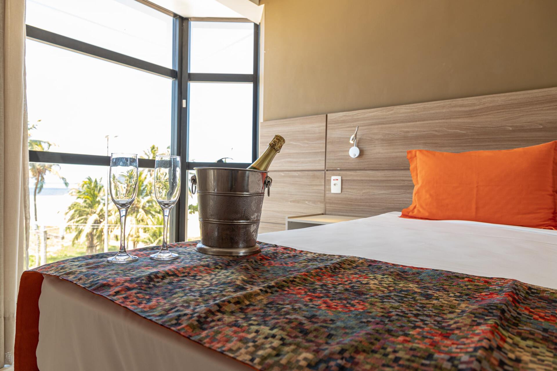 Quarto moderno com cama, almofadas laranja e portas de varanda com vista para a vegetação.
