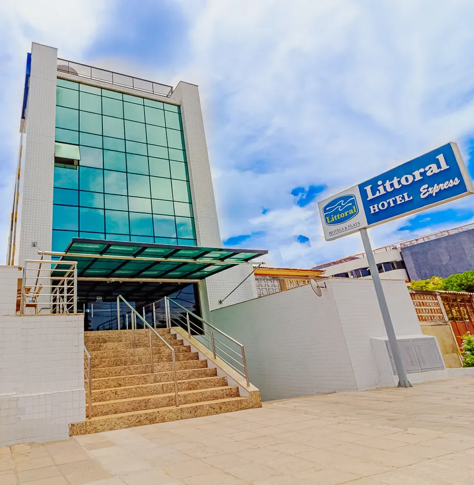 Entrada moderna do hotel, com fachada de vidro azul, escadas e uma placa com os dizeres “Litoral Hotel Capanema”.