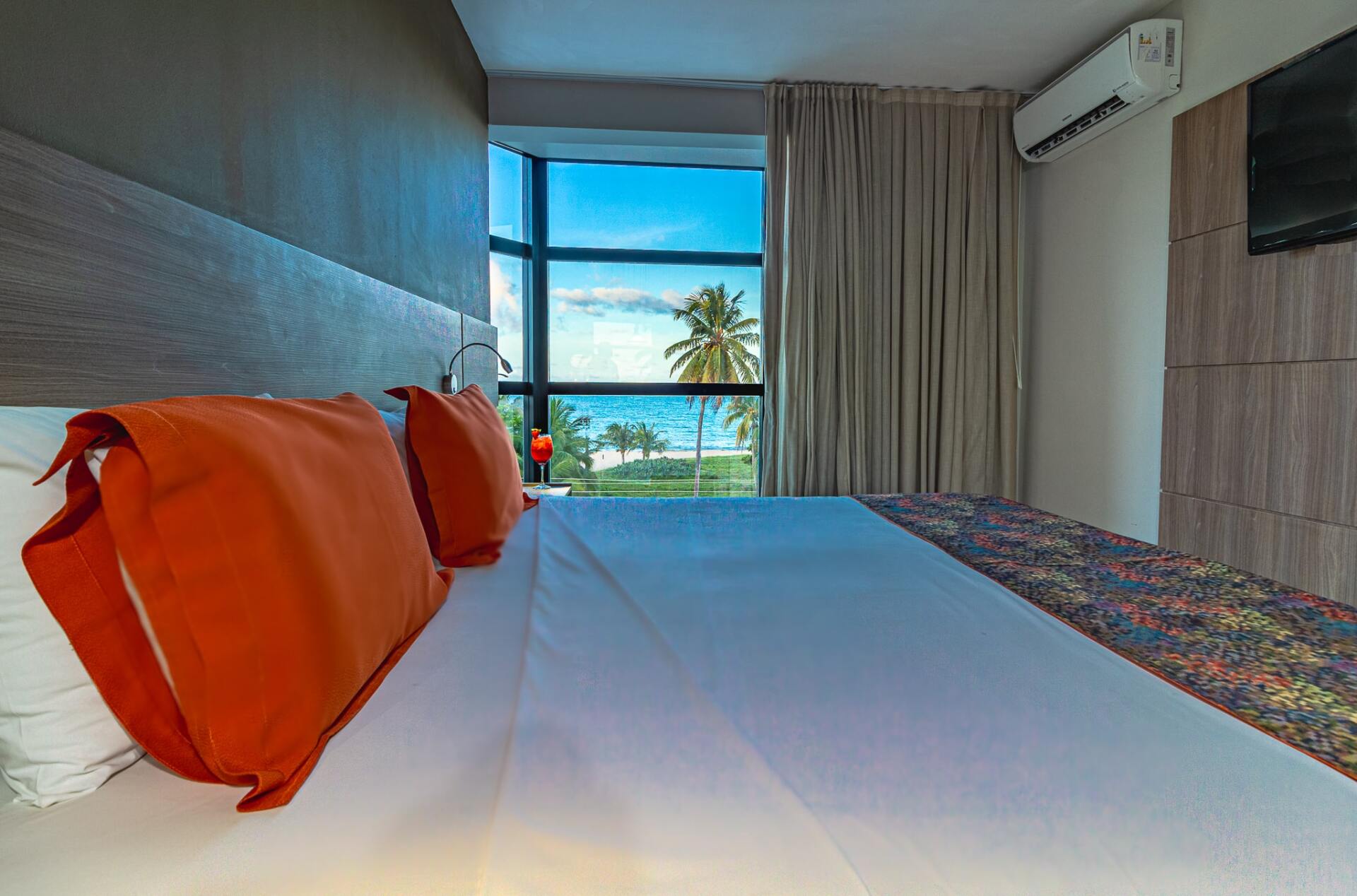 Quarto de hotel com duas camas de solteiro, travesseiros laranja e uma grande janela com vista para palmeiras e o oceano.