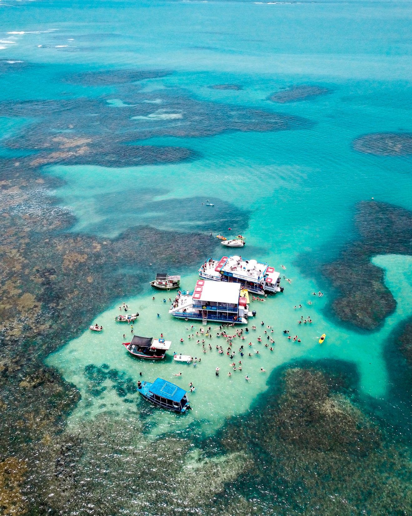Vista aérea de pessoas reunidas em um pequeno banco de areia em águas rasas de um azul turquesa brilhante, perto de recifes de coral.