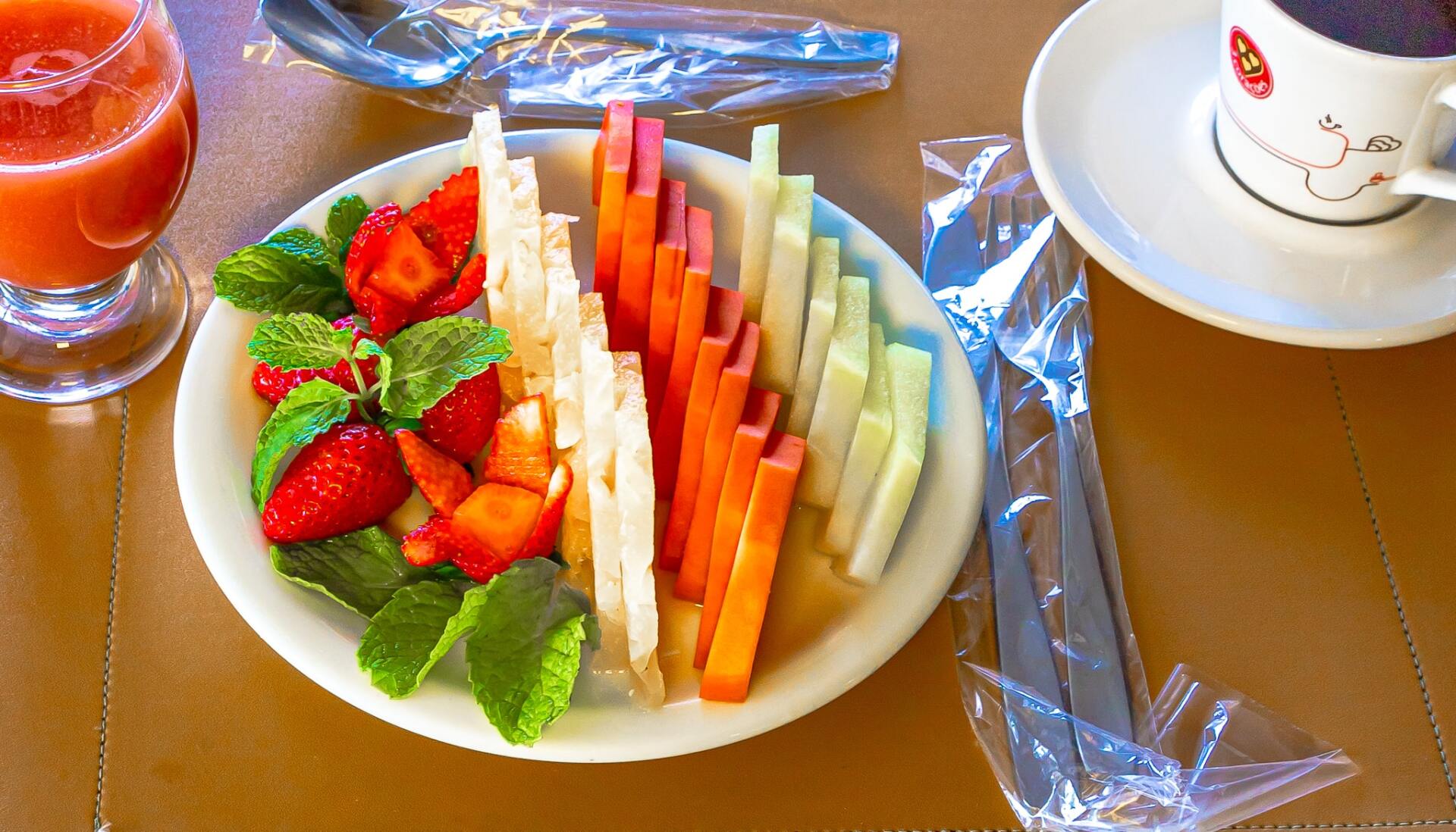 Prato de frutas com morangos, kiwi e fatias de melão sobre uma mesa, ao lado de uma bebida e uma xícara de café.