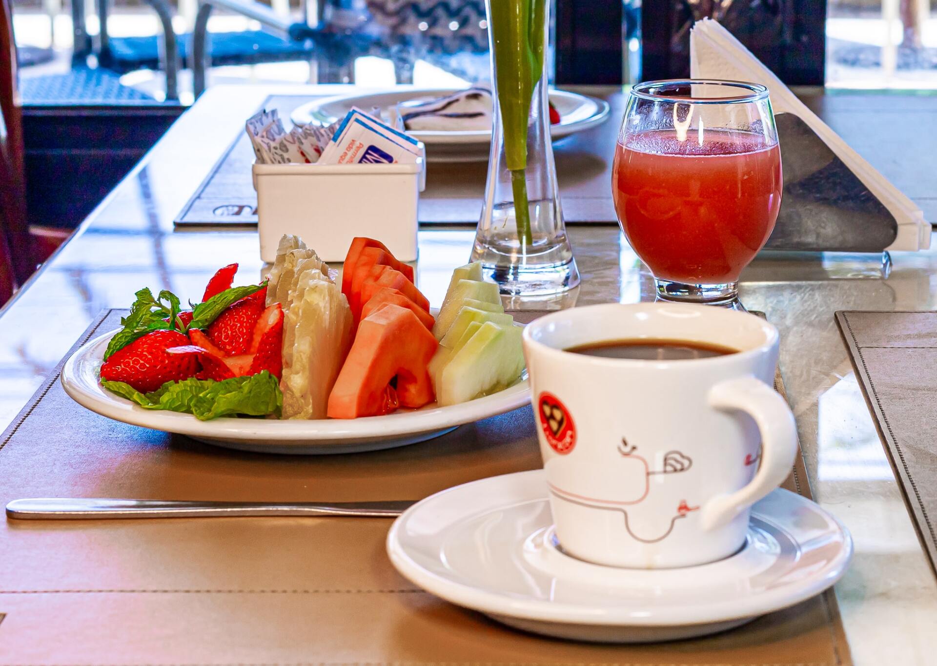 Bandeja de café da manhã com frutas, café e suco sobre uma mesa perto da janela.