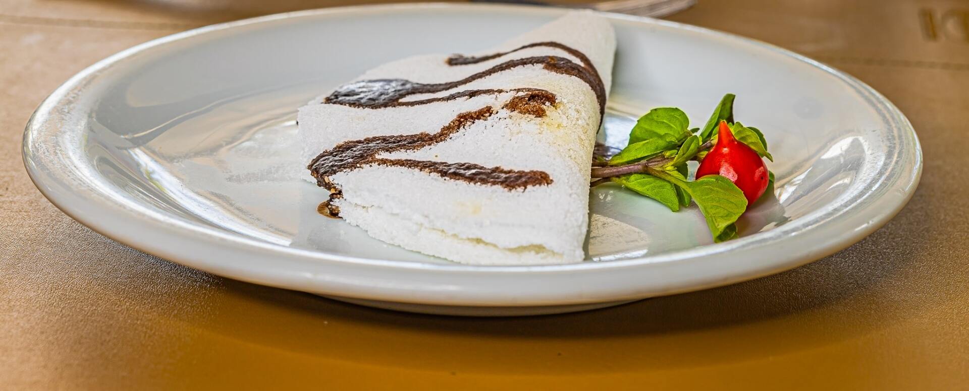 Fatia de bolo com cobertura branca e calda de chocolate, servida em um prato e decorada com morangos e folhas de hortelã.