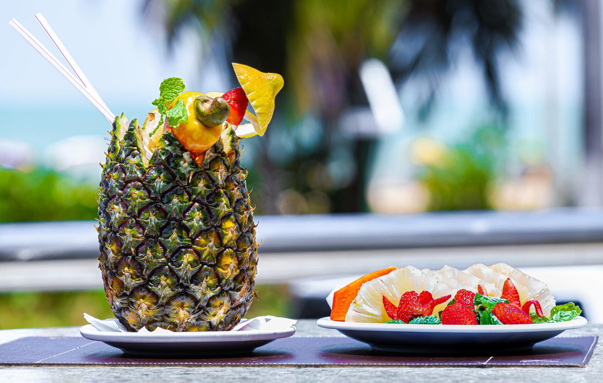 Coquetel de abacaxi com guarnição de frutas ao lado de um prato de sobremesa em uma mesa ao ar livre.