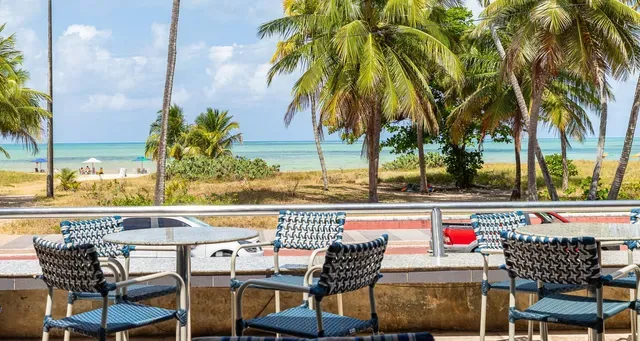 Mesas de café ao ar livre de frente para uma praia tropical com palmeiras e água turquesa.