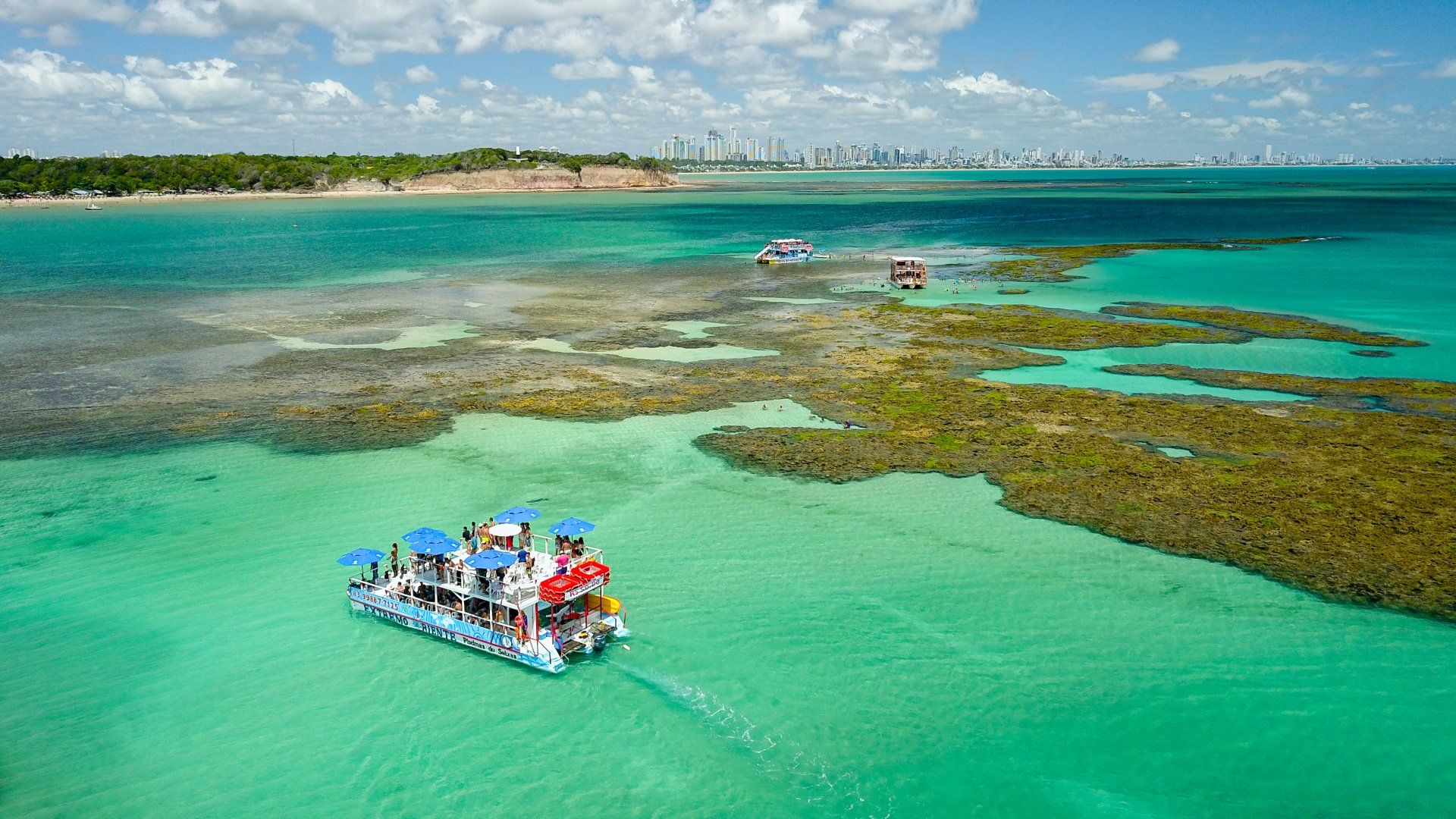 Catamarã com passageiros coloridos flutuando em águas turquesas perto de recifes de coral e uma costa distante.