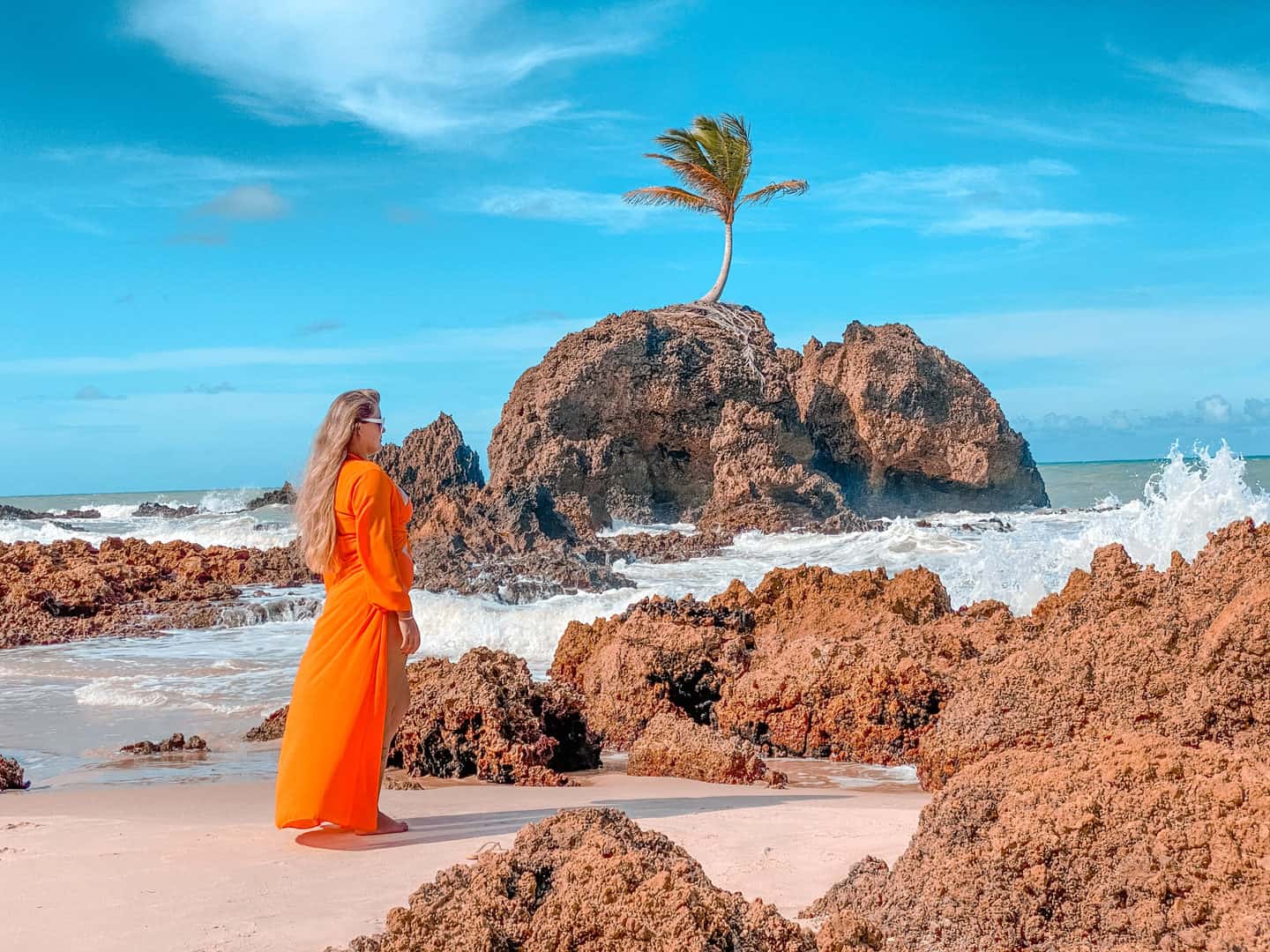 Pessoa vestida de laranja em uma praia rochosa ao lado das ondas e de uma pequena ilha com palmeiras.
