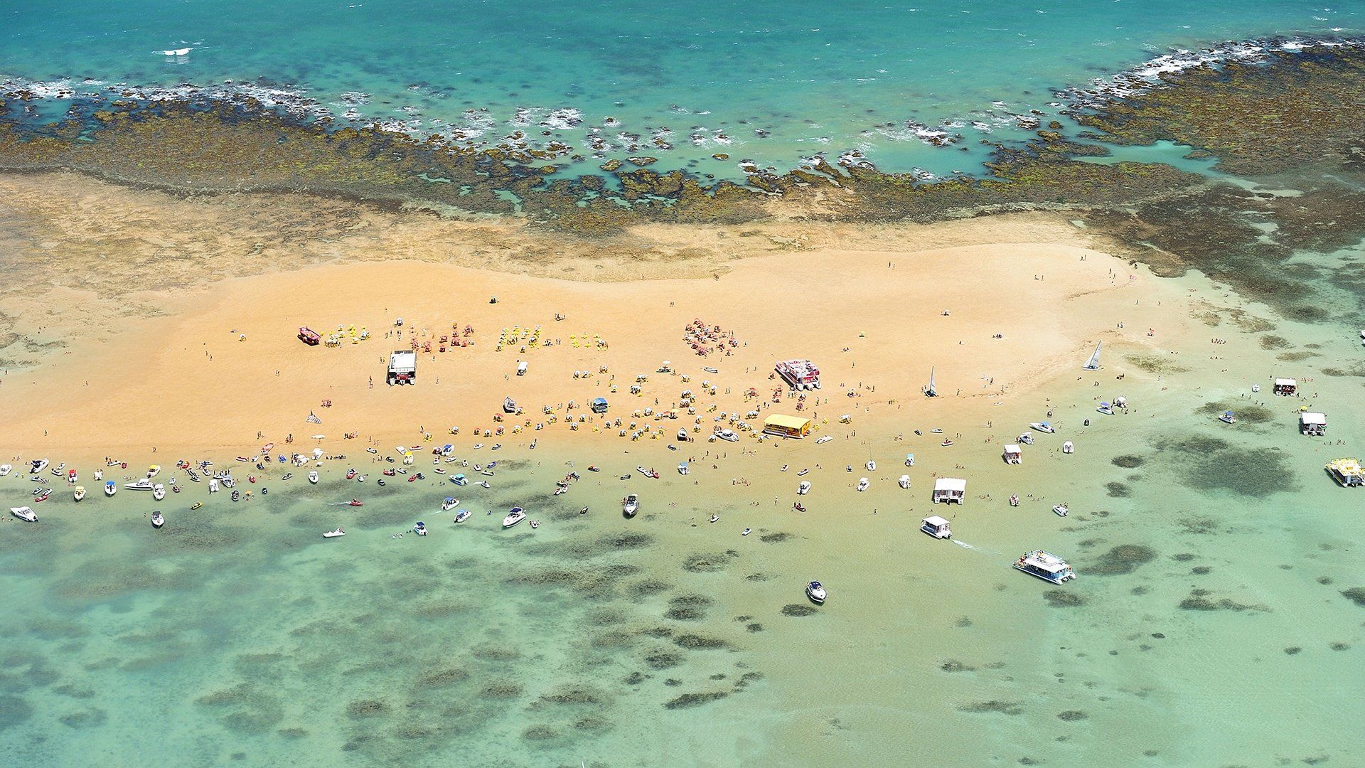 Vista aérea de uma praia de areia com água turquesa e pessoas dispersas perto da costa.