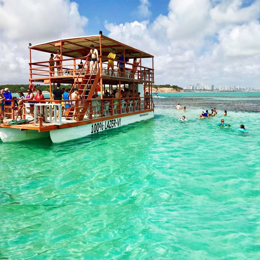 Barco turístico com pessoas perto de banhistas em águas azul-turquesa brilhantes em uma praia ensolarada.