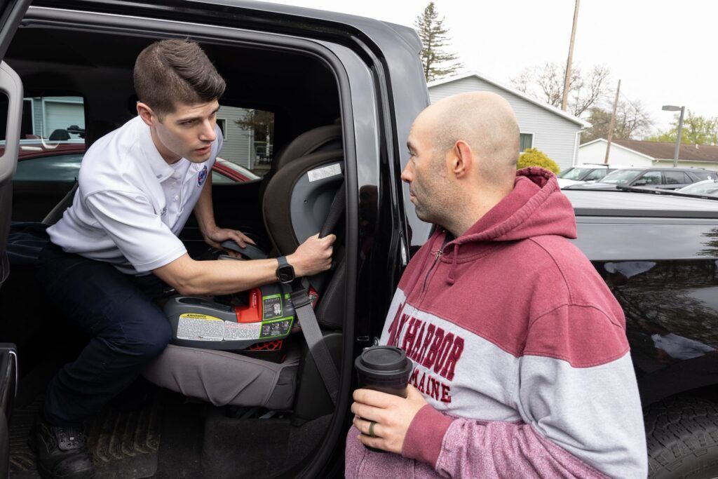 Man loading supplies into a car trunk while another man holds a coffee cup outside in a parking lot