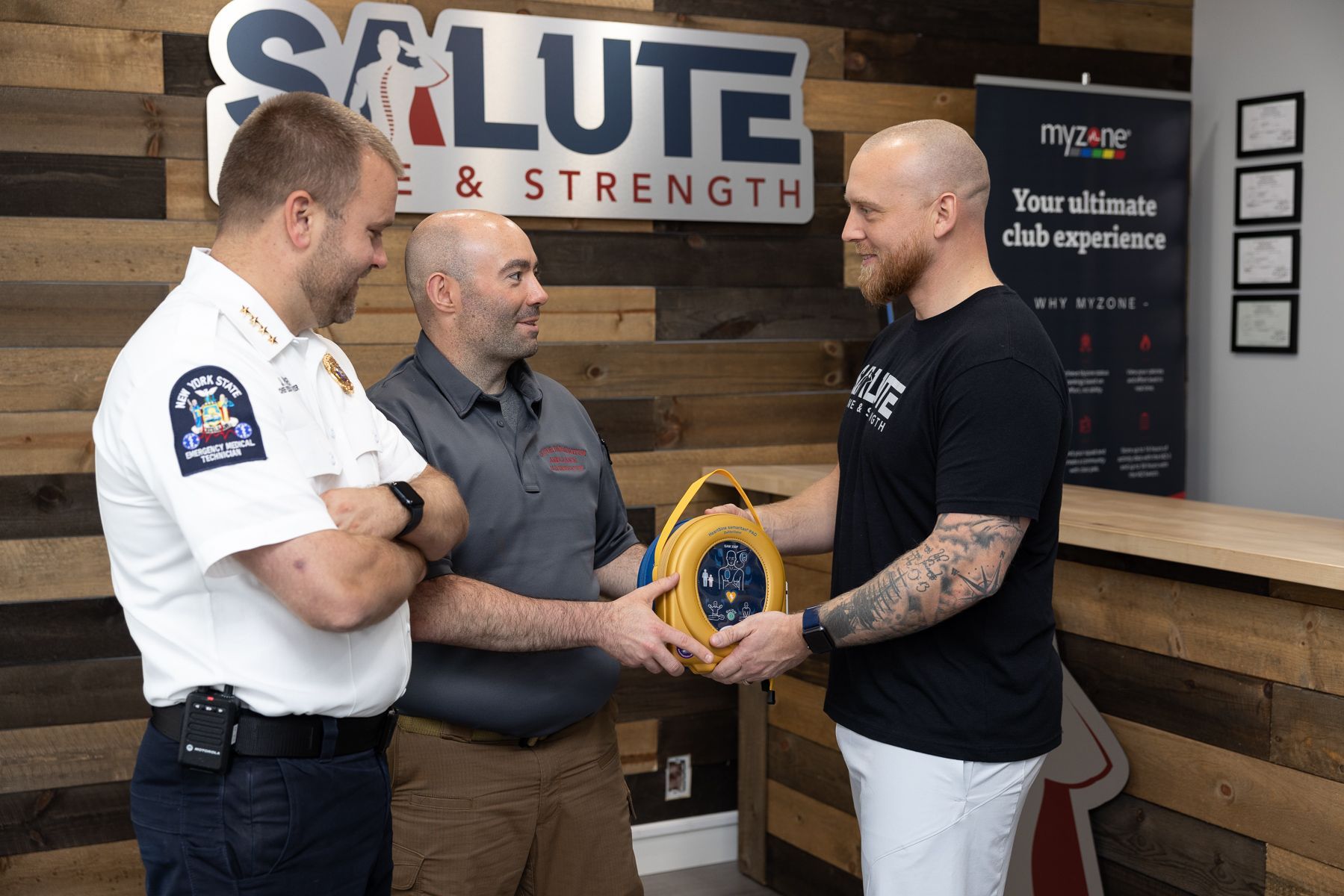Three men in a gym shake hands beside a yellow award plaque and a “Salute & Strength” sign.