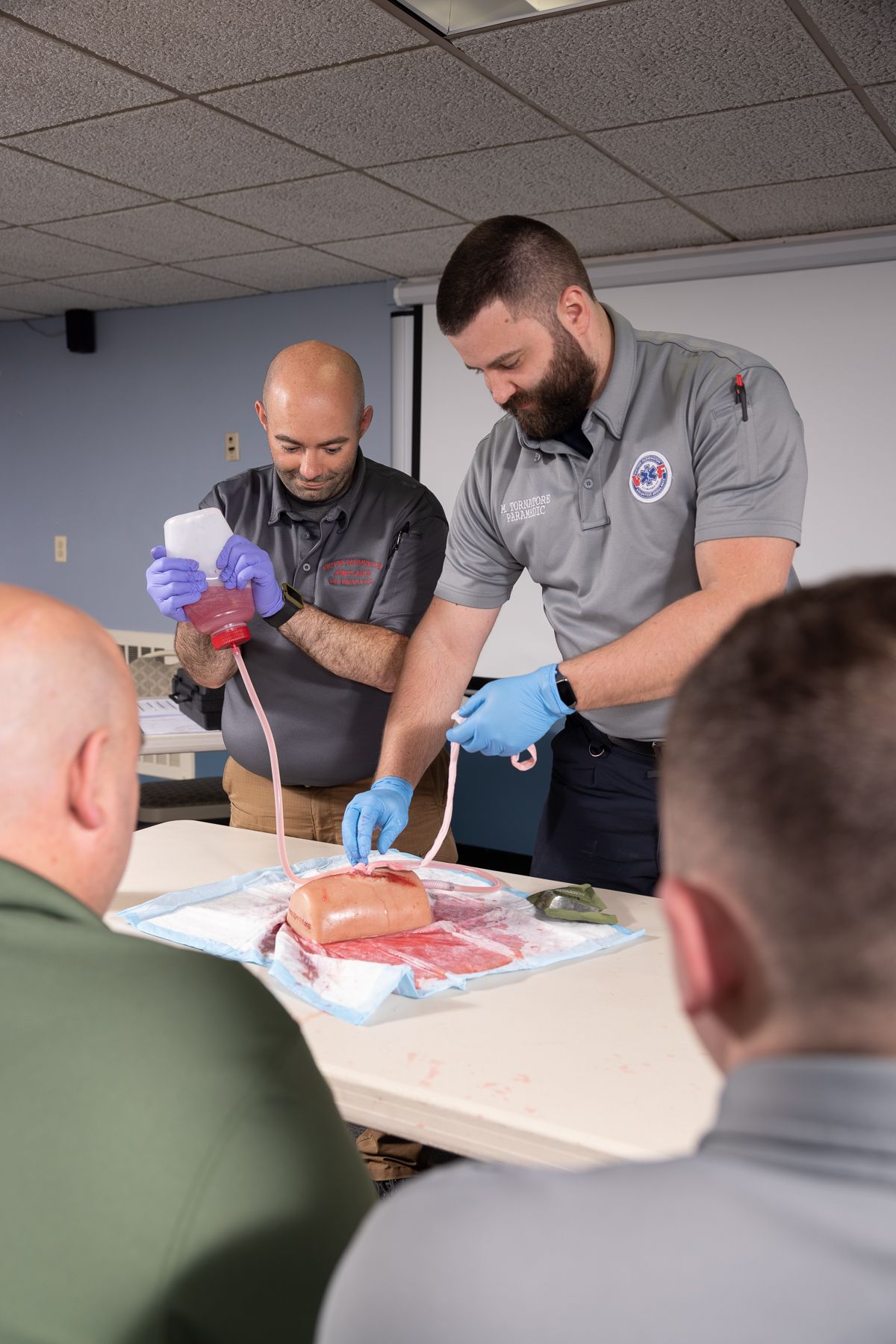 Two men in gray polos demonstrate medical procedure on a model organ in a classroom.