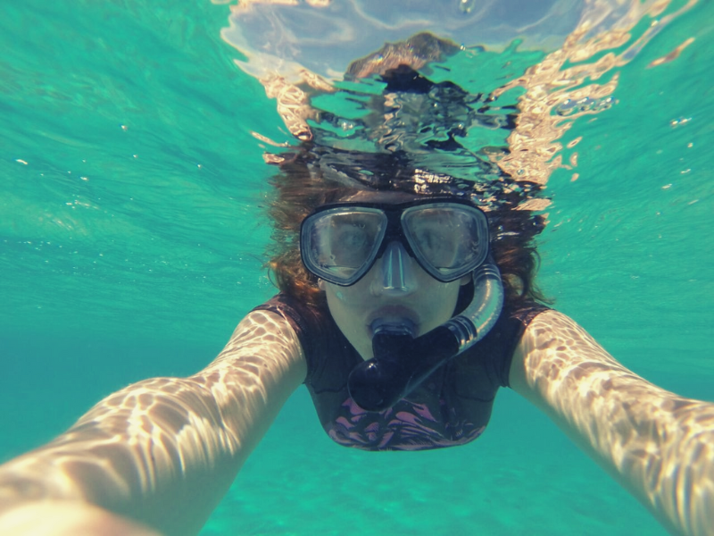 Woman snorkeling underwater, taking a selfie. Wearing mask, snorkel, and black shirt in clear turquoise water.