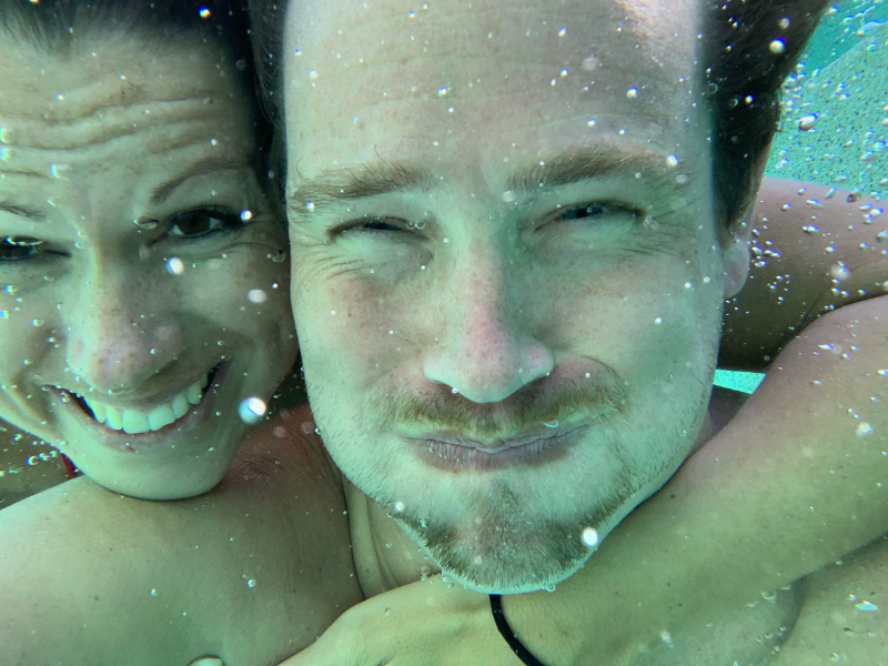 Couple underwater, smiling at the camera. The woman hugs the man. Blue water.