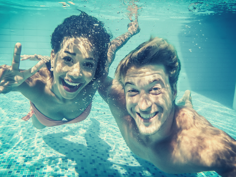 Two people smiling underwater in a pool, one giving a peace sign, clear blue water.