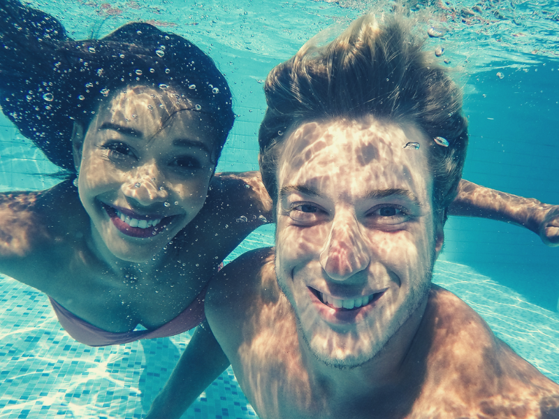 Couple underwater, smiling at the camera. Woman with dark hair, man with blond hair, both in a pool.