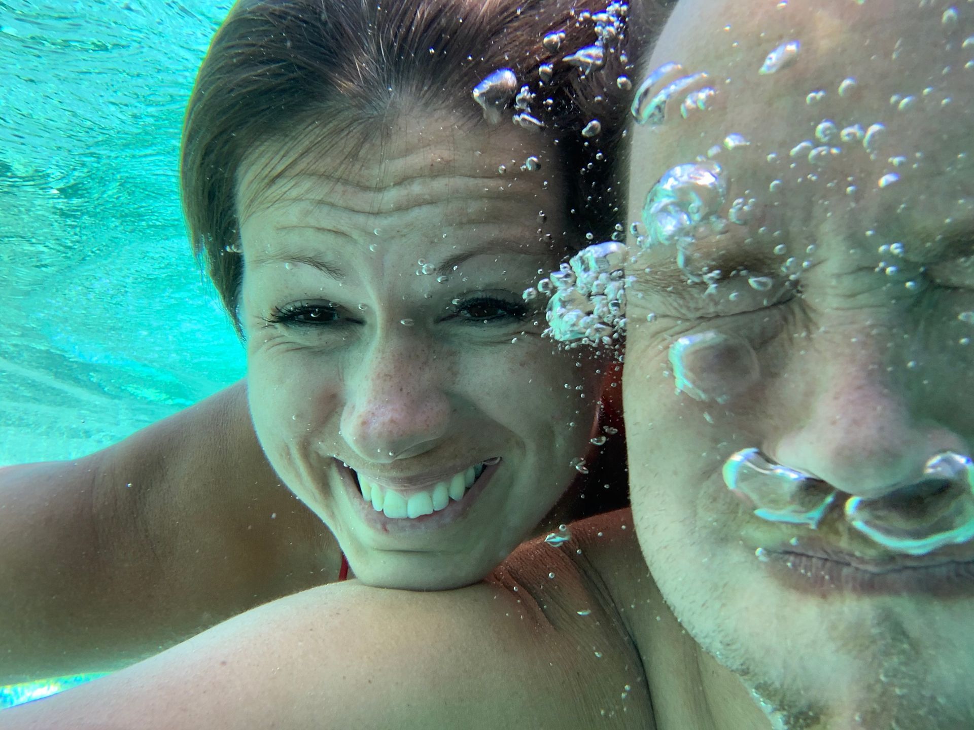 Woman smiles underwater; man's face closed, bubbles.