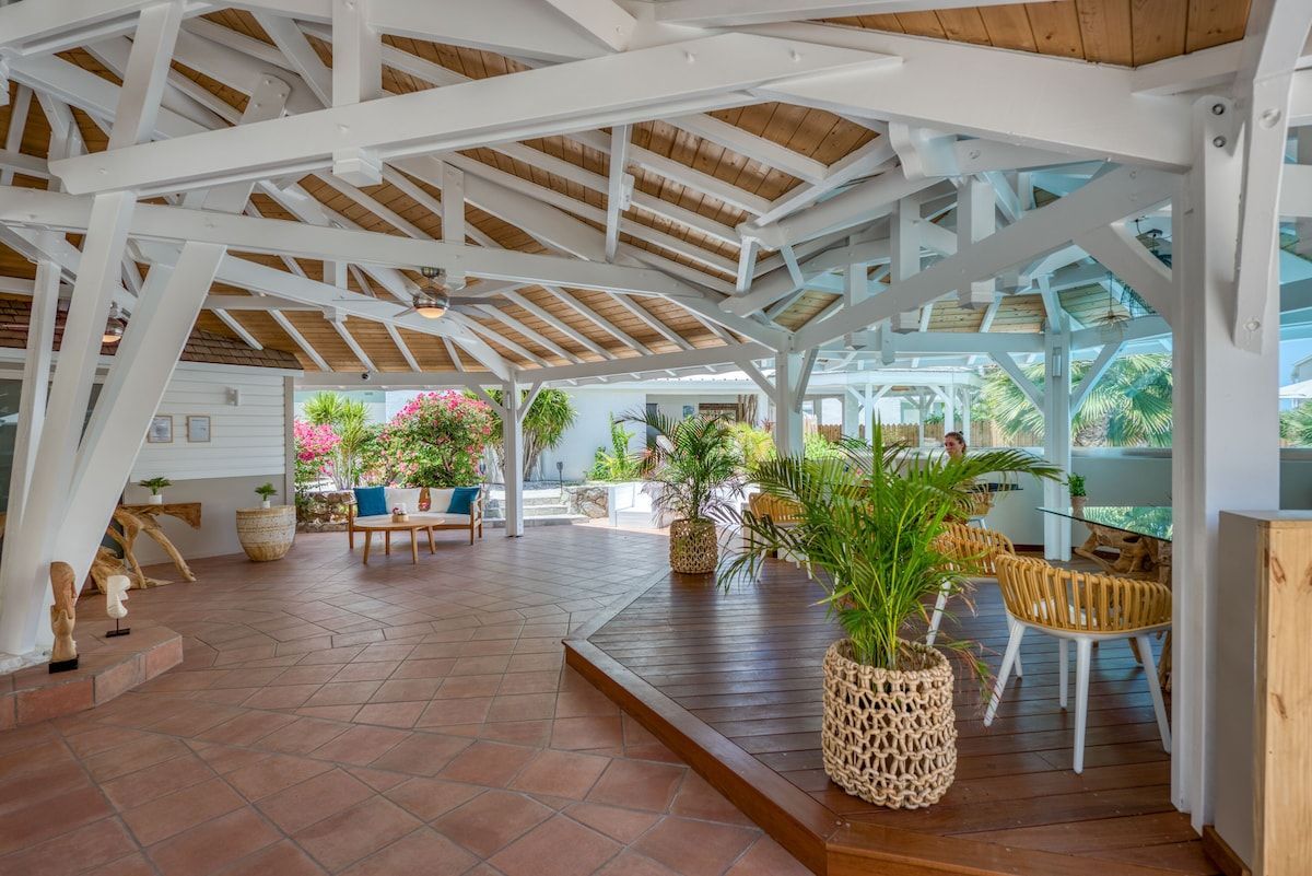 Open-air white-beamed patio with red tile floors, wood deck, plants, and seating overlooking lush garden.