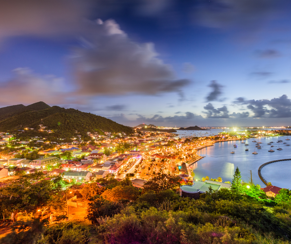 Coastal town at dusk with bright lights, buildings, harbor, boats, and hills.