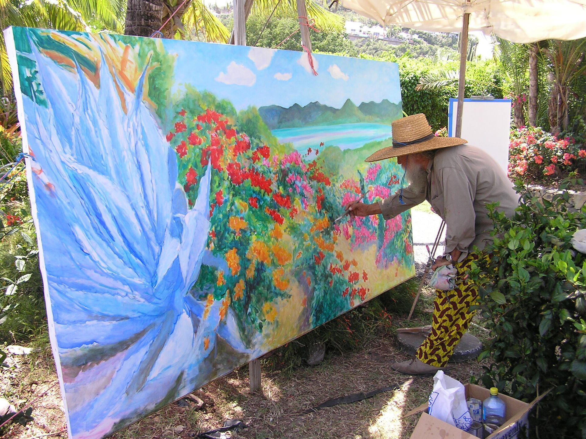 Artist painting a large outdoor mural of a colorful garden, wearing a straw hat and patterned pants.