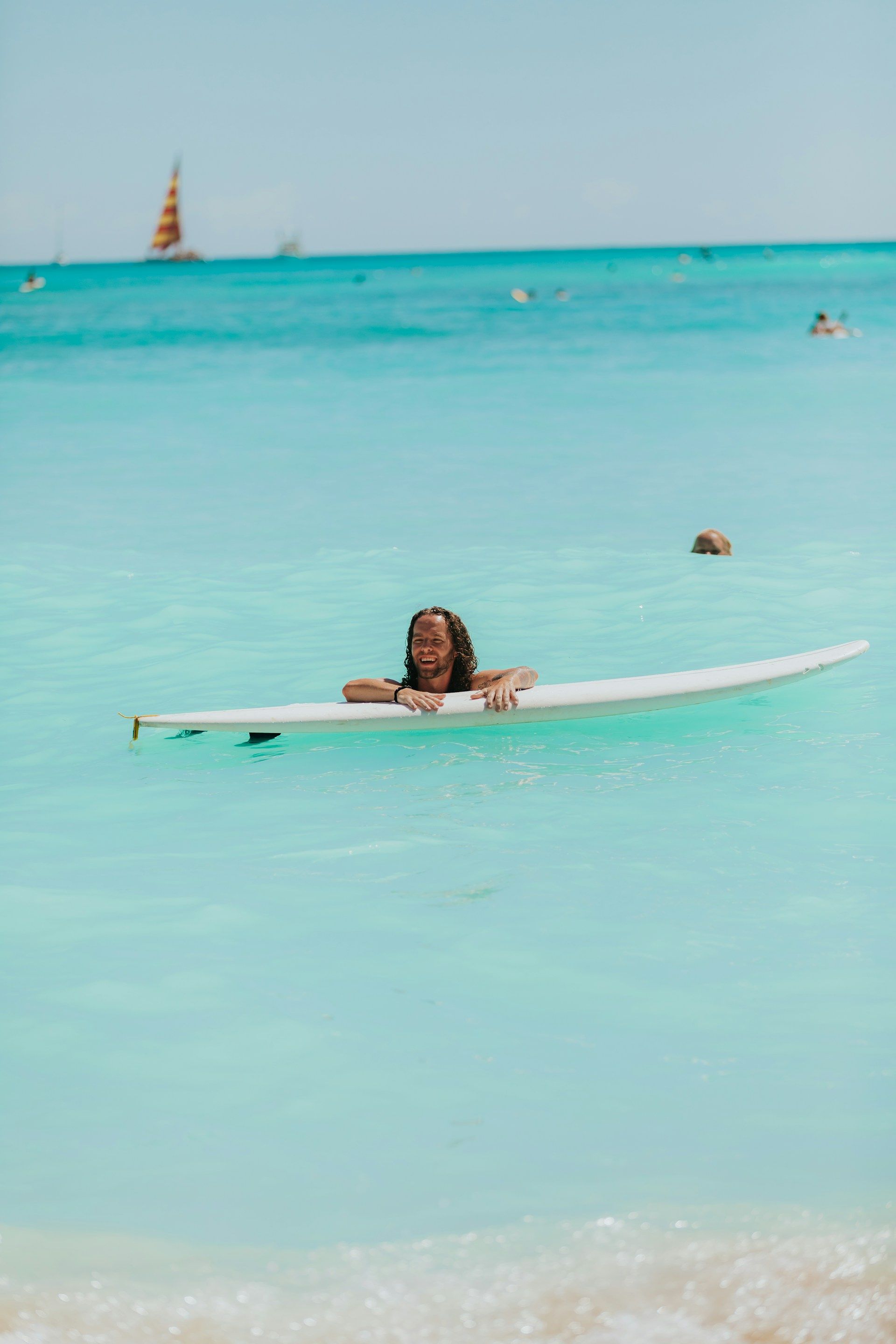 Person on a surfboard in clear turquoise water, near a beach with a sailboat in the distance.