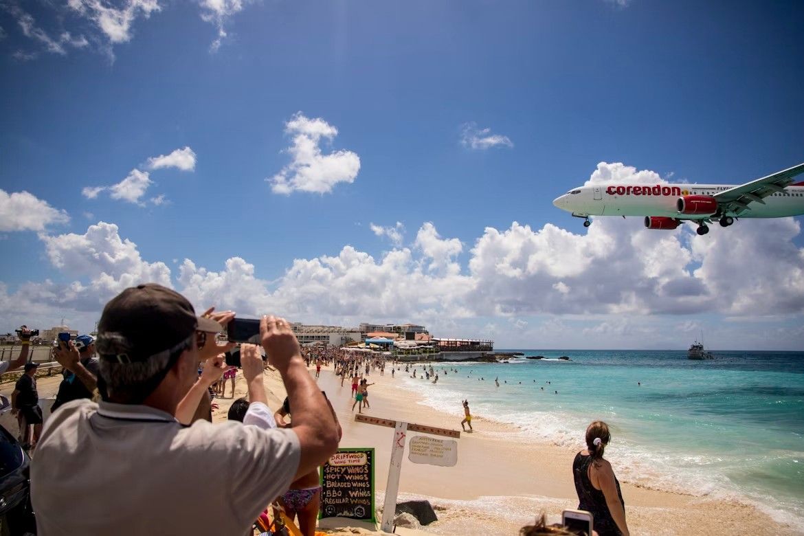 People watch an airplane land over Maho Beach, St. Maarten, under a partly cloudy sky.