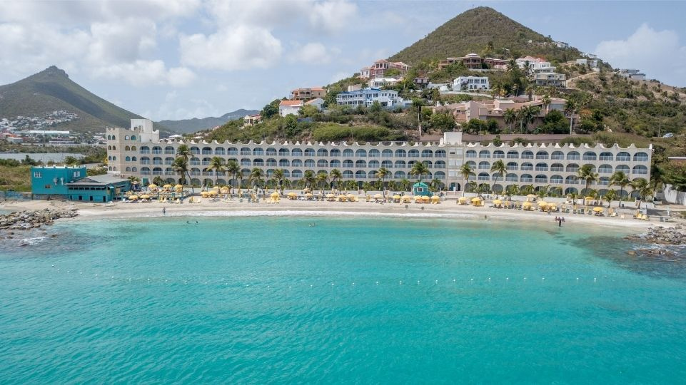 Beachfront hotel on a turquoise sea with a mountain backdrop and a partly cloudy sky.