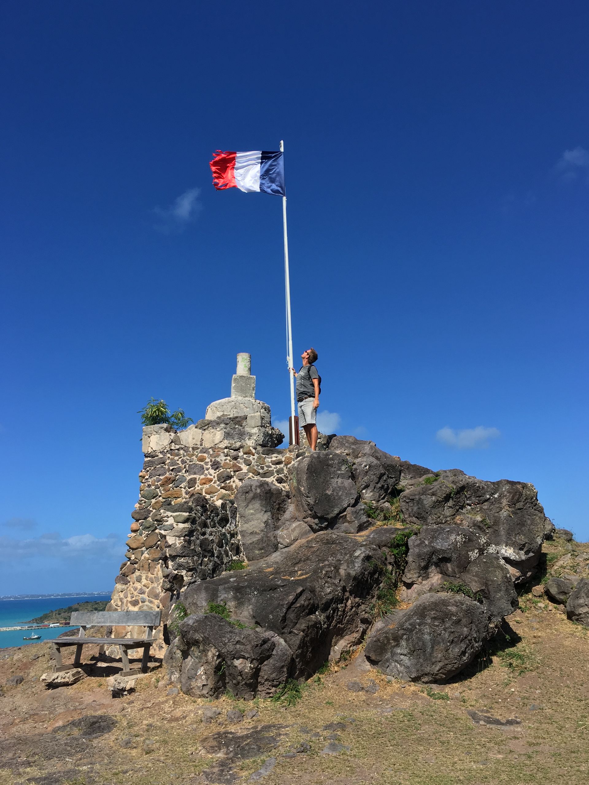 Man standing on rocky outcrop with French flag against a blue sky.