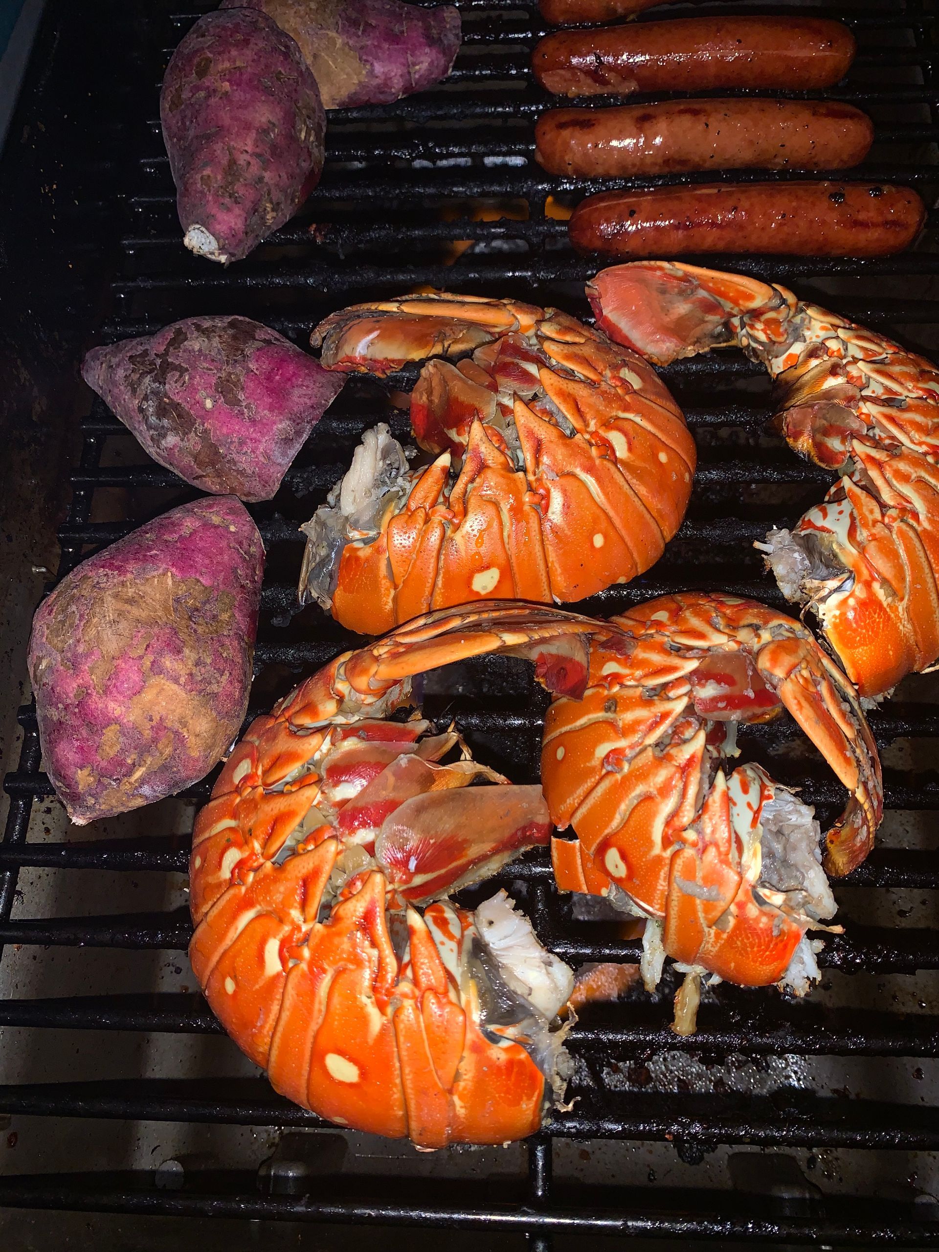 Lobster tails, sausages, and sweet potatoes grilling on a barbecue.